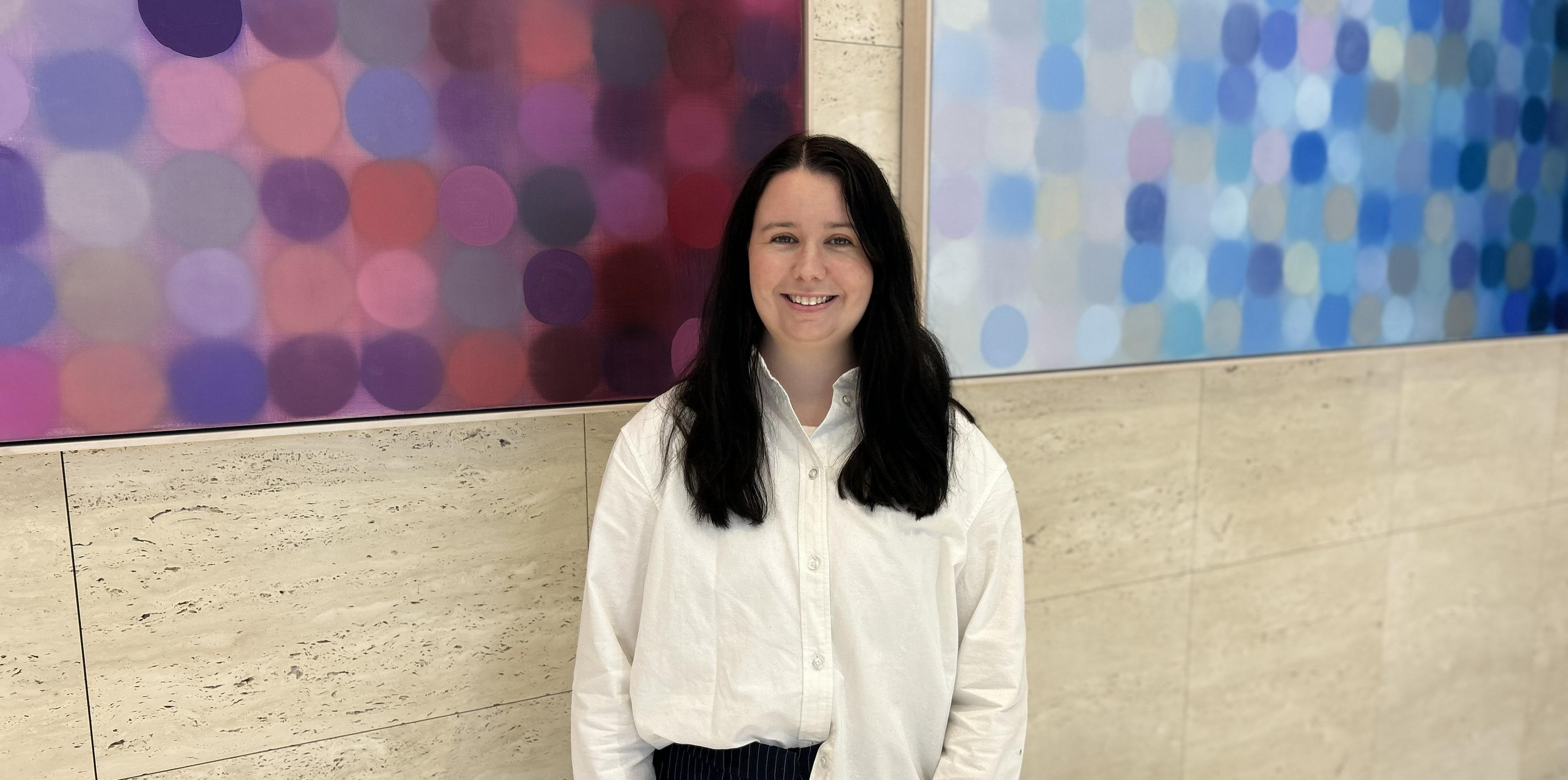 A woman with long dark hair, wearing a white shirt, stands smiling in front of colorful abstract art on a stone wall background.